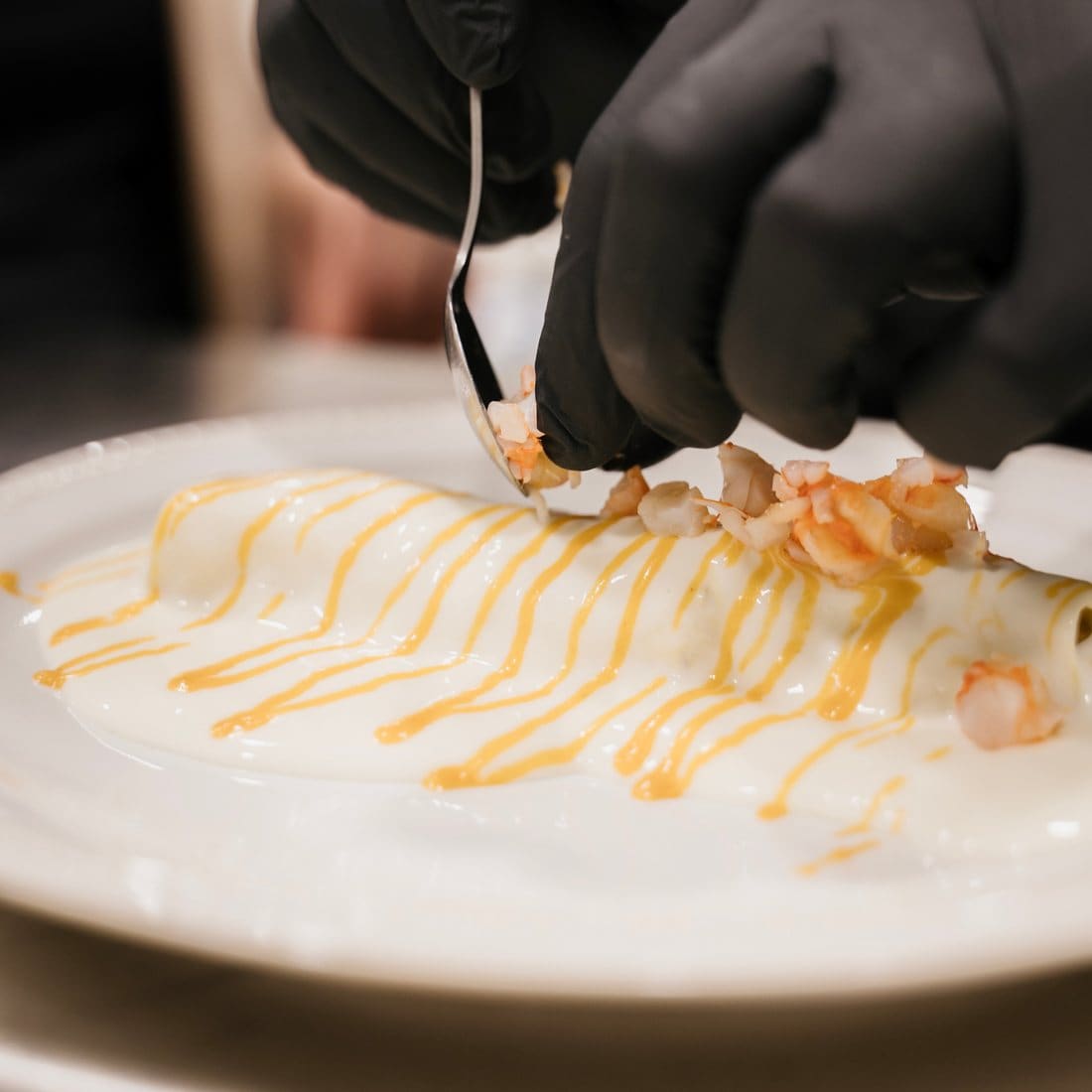 Mans amb guants acabant l’emplatat d’un plat amb beixamel, fils de salsa i trossets de gamba