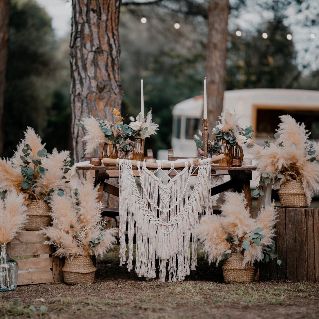 Racó decorat amb pampes, macramé i espelmes davant d’una caravana vintage, en un esdeveniment entre pins