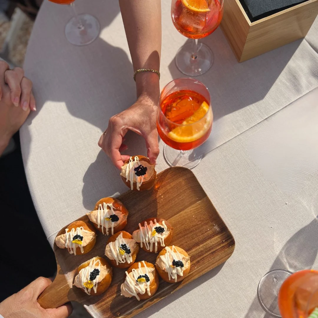 Manos cogiendo una lionesa con caviar de una tabla, con cócteles naranjas