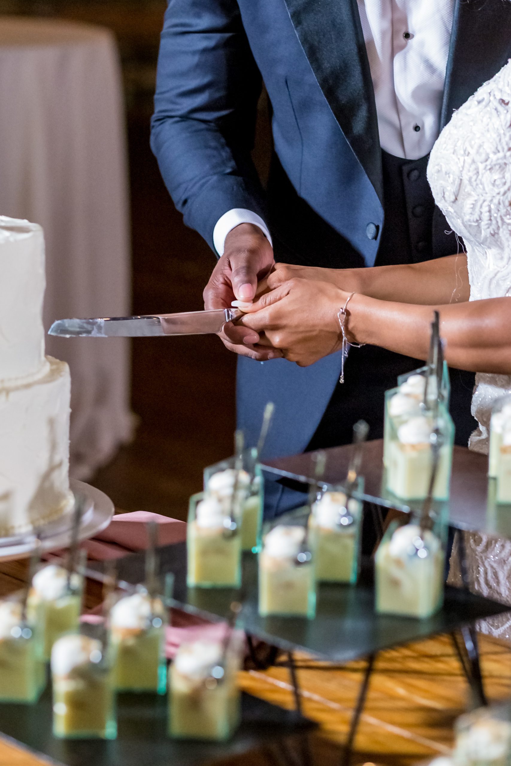 Manos de los novios cortando una tarta blanca con copas de postre delante