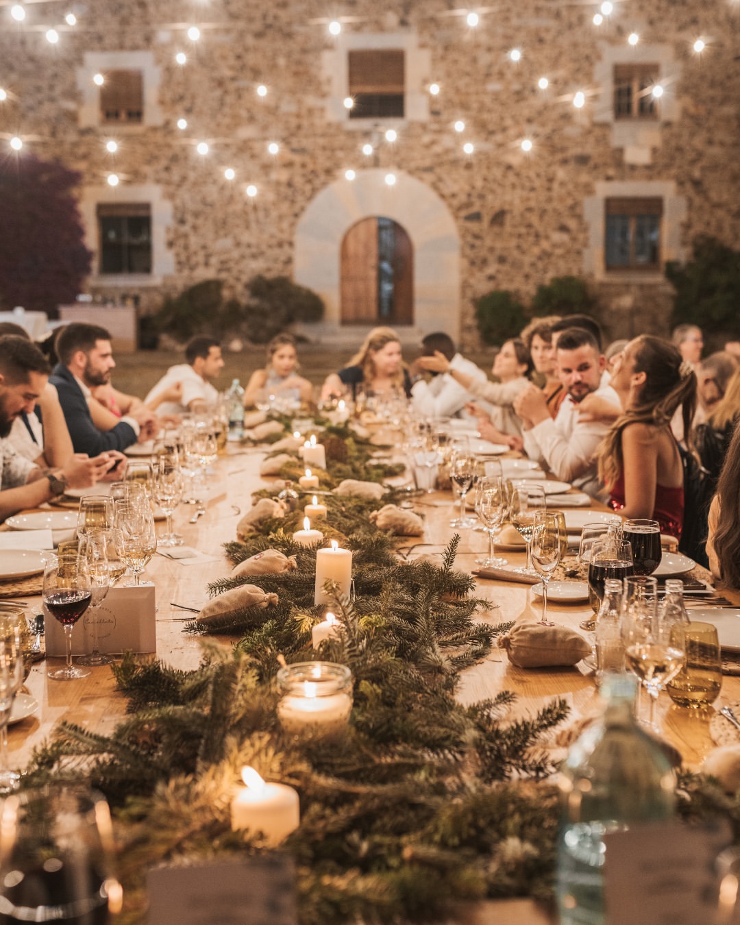 Convidats sopant en una taula llarga davant una masia de pedra il·luminada