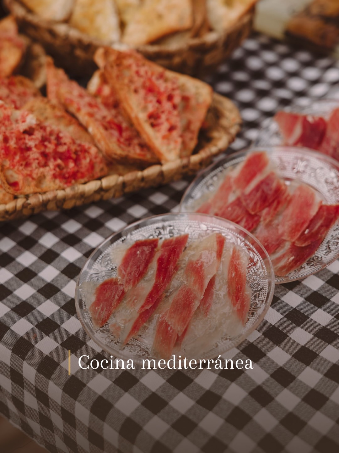 Cesta de pan con tomate y platos de jamón cortado sobre mantel de cuadros